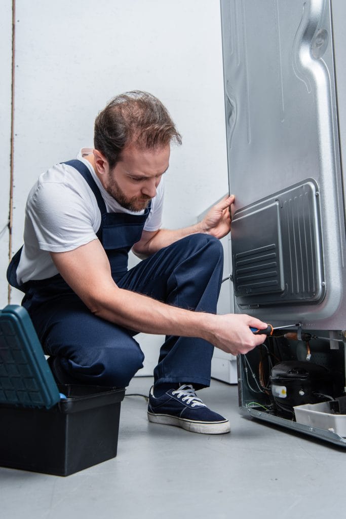 A repairman in overalls is crouching and using a screwdriver to fix the back of a refrigerator during an appliance repair and installation service.