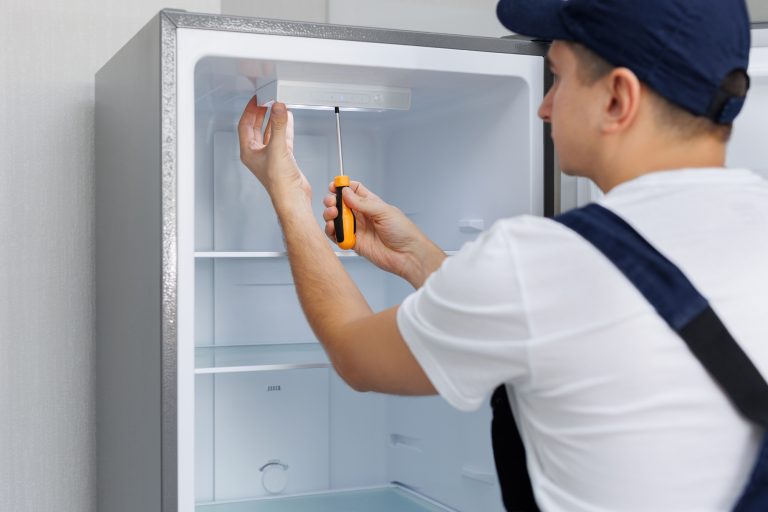 man in a uniform repairs the light in the refrigerator with a screwdriver replacing the light bulb