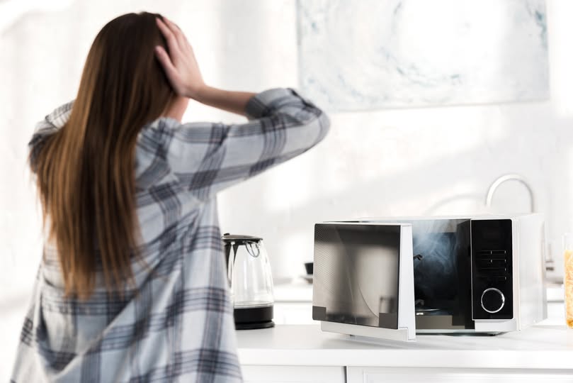 back view of shocked woman looking at broken microwave in kitchen