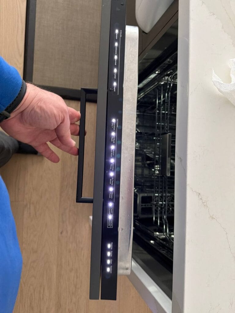 A person’s hand holds the handle of an open dishwasher, ready for appliance repair and installation, showing its illuminated control panel and empty racks inside.