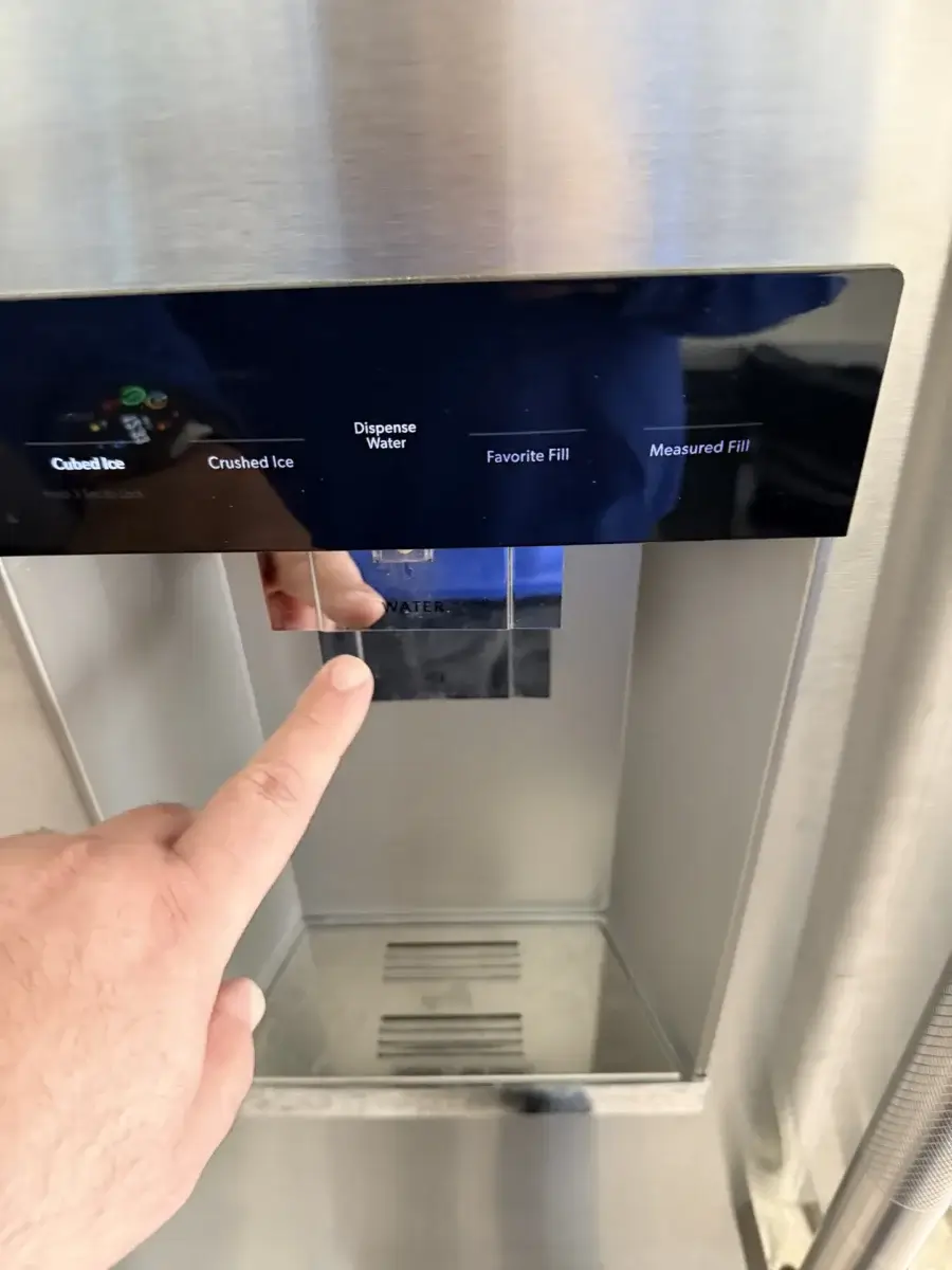 Hand pressing the water dispenser on a stainless steel refrigerator, fed by a supply line that can leak
